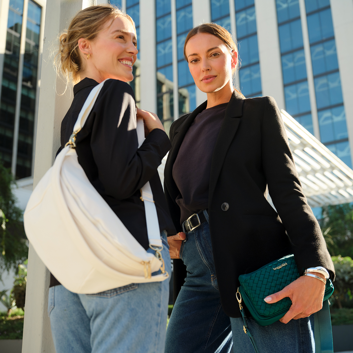 Two women standing outdoors in front of a modern glass building, both wearing casual clothing and carrying baggallini handbags, looking toward the camera.